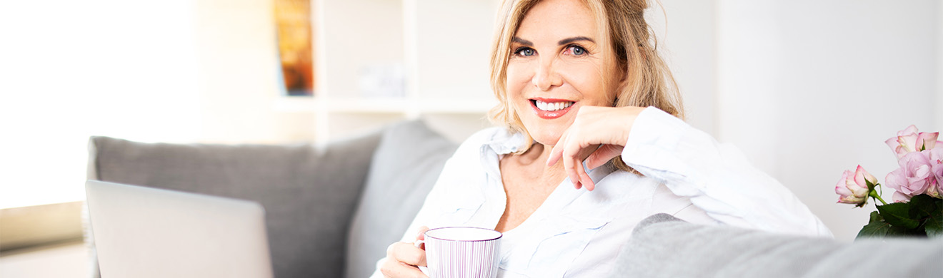 Sympathisch lächelnde Frau sitzt entspannt mit Laptop und Kaffeetasse auf der Couch in heller Wohnung.