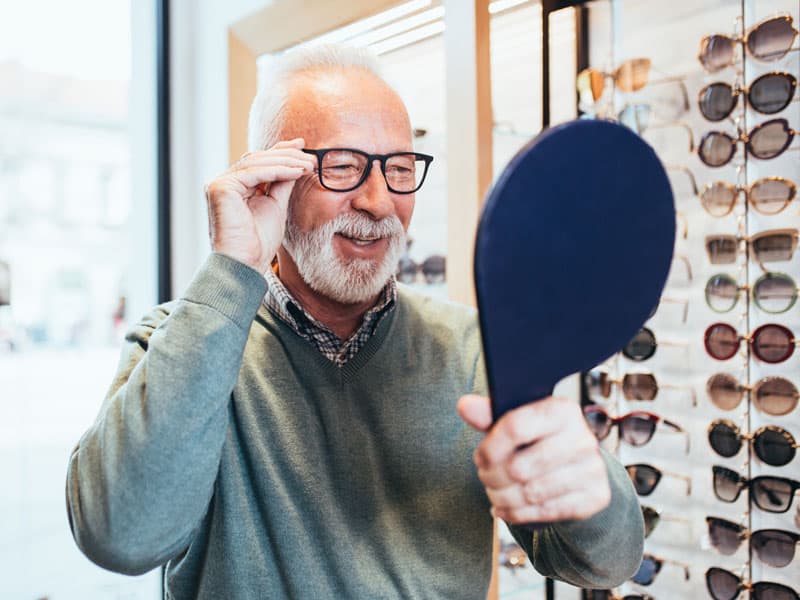Älterer Mann probiert neue Gleitsichtbrille vor Spiegel an, steht vor Brillengestell-Auswahl im Optikergeschäft in Flensburg.