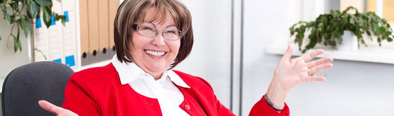 Ältere Frau mit Gleitsichtbrille lacht freundlich im Büro, trägt roten Blazer und sitzt am Schreibtisch.