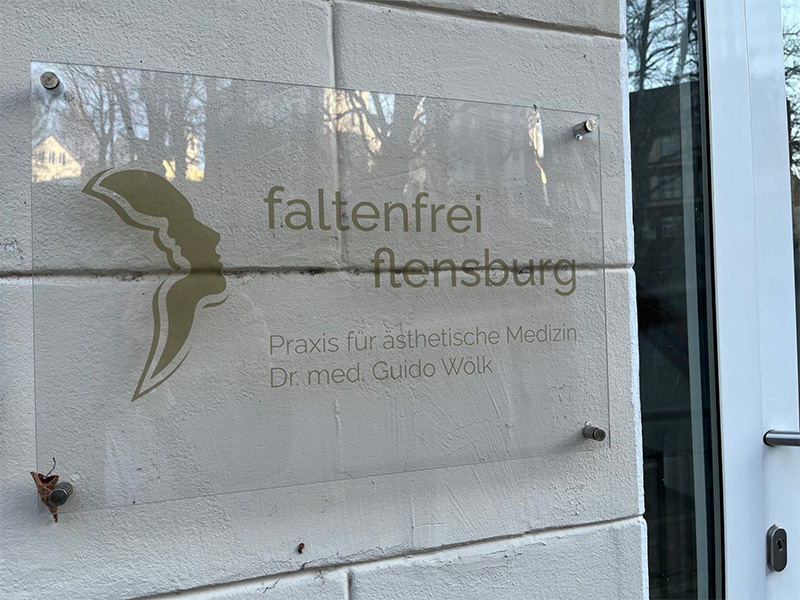 Praxisschild auf durchsichtigem Plexiglas montiert an Hauswand neben Eingang - sauber und professionell.