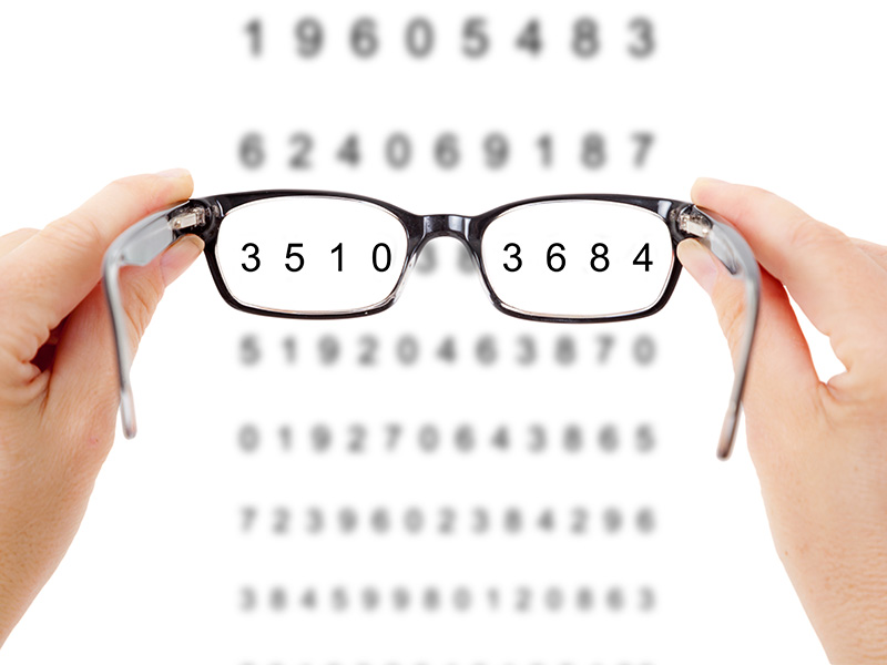 Eine Hand hält eine Brille mit schwarzem Rahmen vor eine Zahlenreihe mit neutralem, unscharfem Hintergrund bei Sehtest in Flensburg.