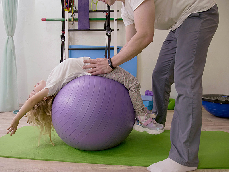 Kind führt Physiotherapieübungen auf Gymnastikball zur Unterstützung der Skoliosebehandlung Kind mit therapeutischer Begleitung in heller Praxisumgebung durch.